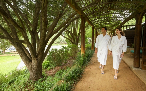 A couple walks under a wooded canopy surrounded by greenery in white Lake Austin branded robes