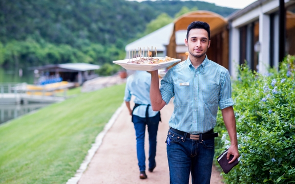A male server in jeans and a nice checkered shirt carries a tray loaded with hors d'hoeuvres along an outdoor path.
