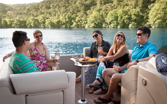 A group of three women and two men out on a boat on the lake. They're all wearing sunglassses, sharing some food, laughing.