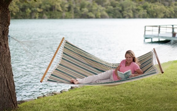 A woman laying in a striped green hammock overlooking the lake while reading a book.