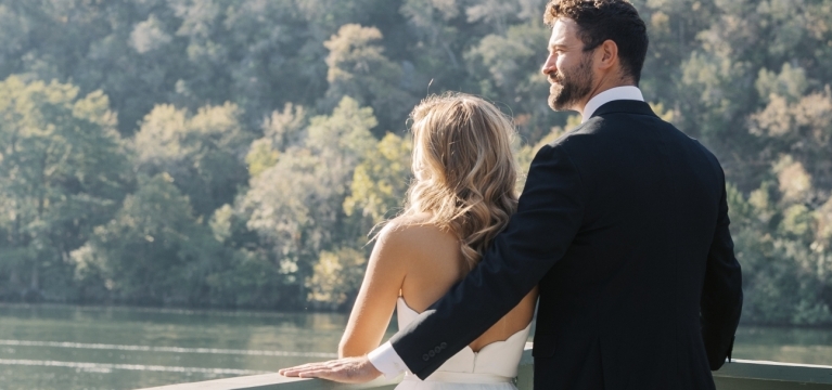Bride and groom standing by the lakeside, facing away from the camera and looking in the same direction