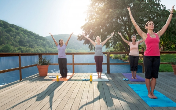 Three women attend a yoga class as their instructor guides them through a series of moves on a deck over the water.