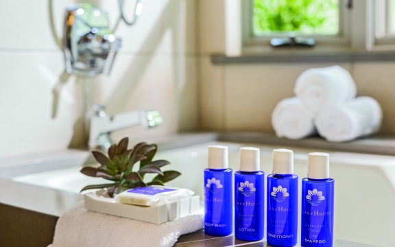 Beautiful blue bottles line the side of a luxurious looking bathtub. There are also towels rolled up and a pretty plant.