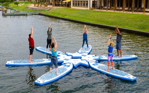 Six people perform yoga on floating boards on a lake with the resort and plenty of greenery in the background.