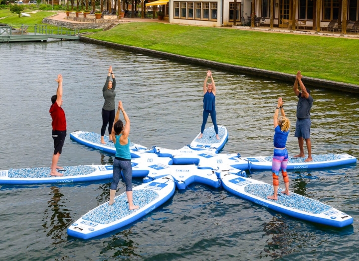 Six people perform yoga on floating boards on a lake with the resort and plenty of greenery in the background.